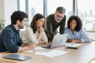 Quatre collègues discutent autour d'une table de travail, regardant un ordinateur portable avec des graphiques, dans un bureau moderne et lumineux avec une vue sur la ville.
