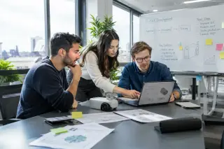 Three young professionals collaborate around a conference table in a modern, sunlight-filled office, with technical documents, a laptop, and a prototype device, evoking an innovative start-up atmosphere.